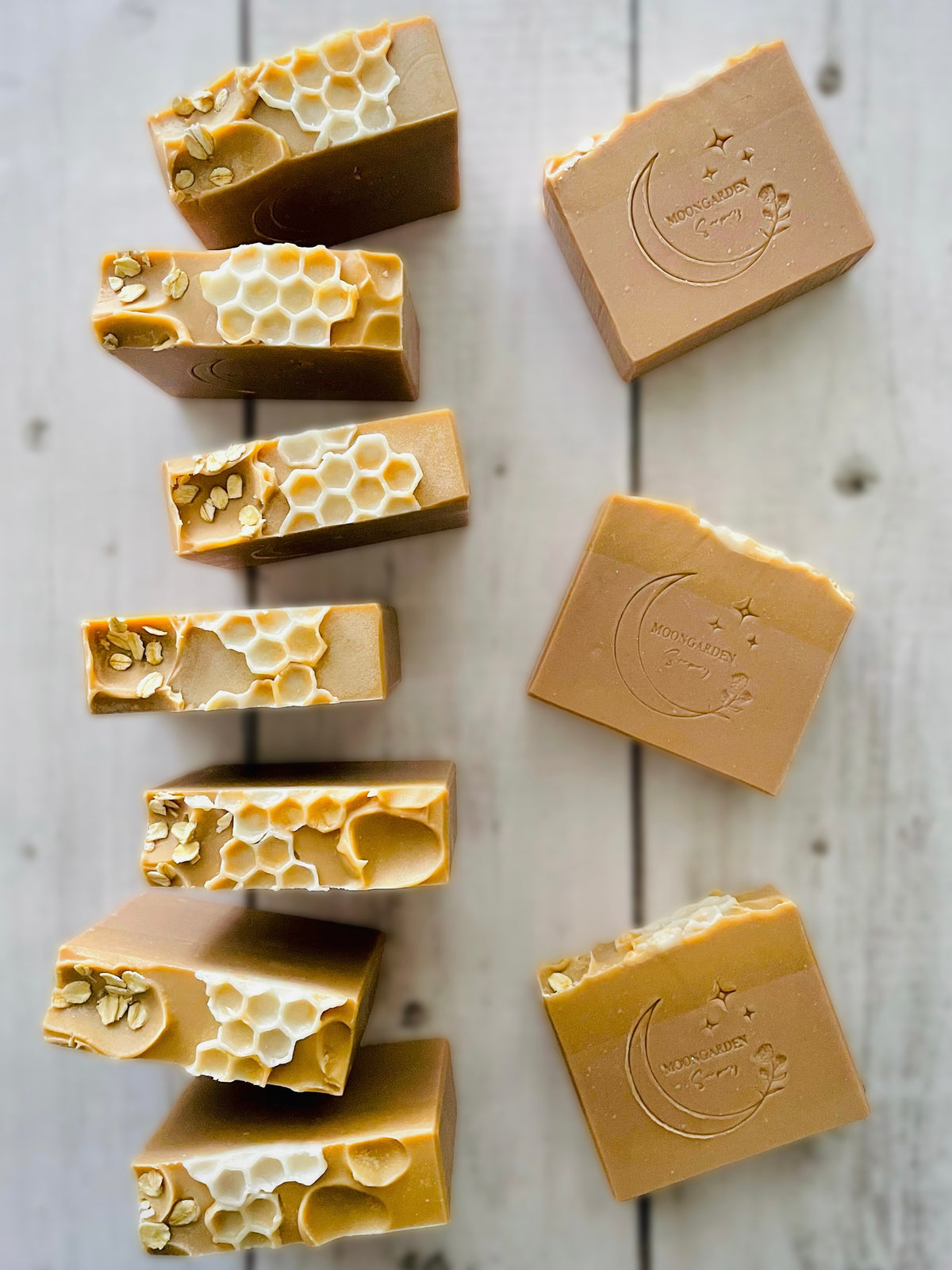 Ten handmade soap bars in tan and gold, topped with oats and honeycomb soap embeds, arranged on a white rustic wooden background.