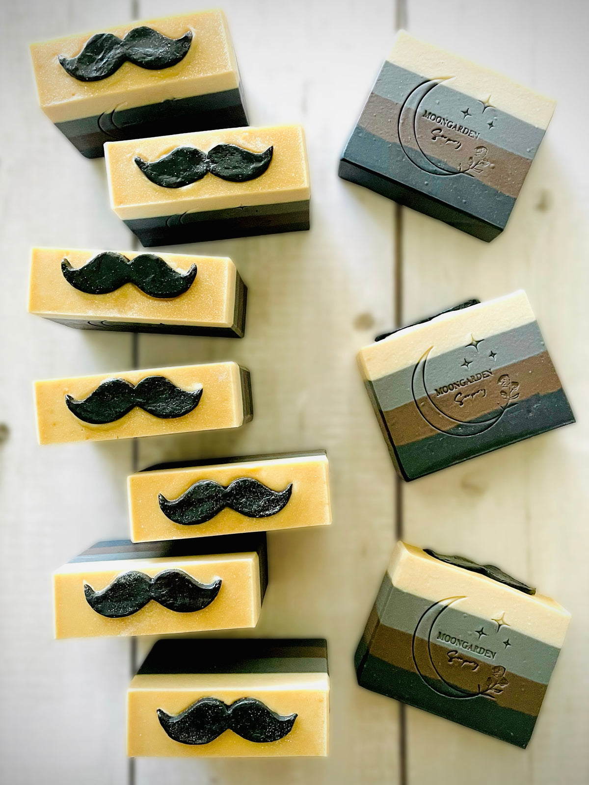 Ten handmade soap bars in black, dark brown, gray, and white, topped with black mustache soap embeds, arranged on a white rustic wooden background.