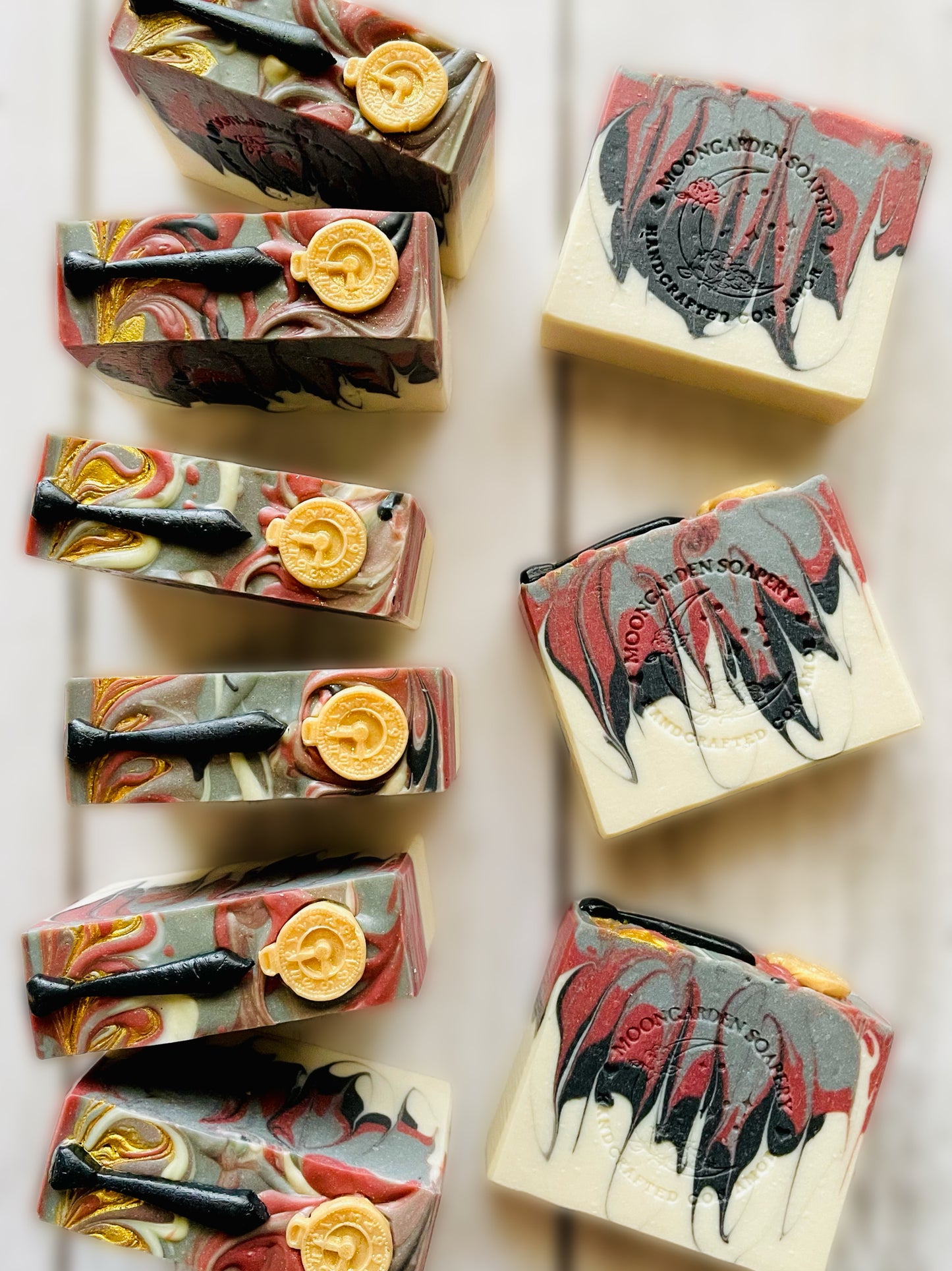 Nine handmade soap bars in white, black, gold, dark red, gray, topped with shirt tie and pocket watch soap embeds, arranged on a white rustic wooden background.