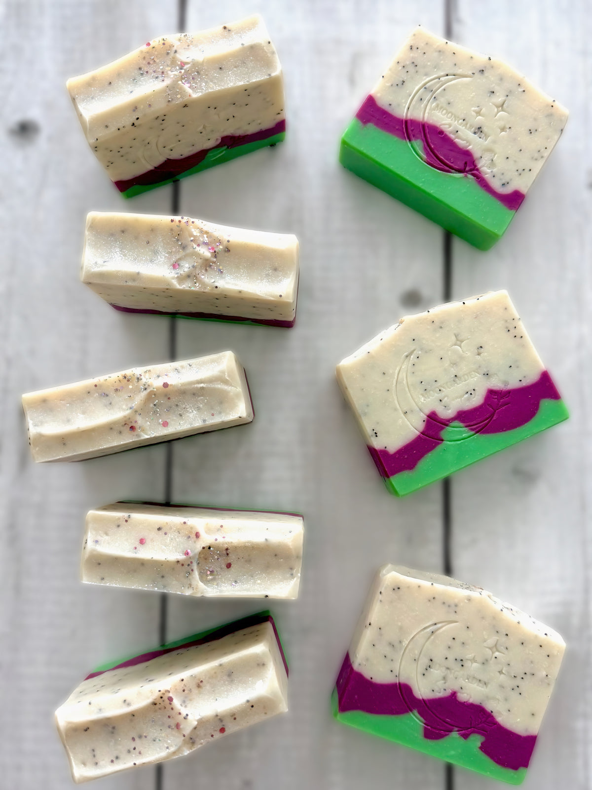 Eight handmade soap bars in neon green, dark pink, and white with poppy seeds, topped with glitter, arranged on a white rustic wooden background.
