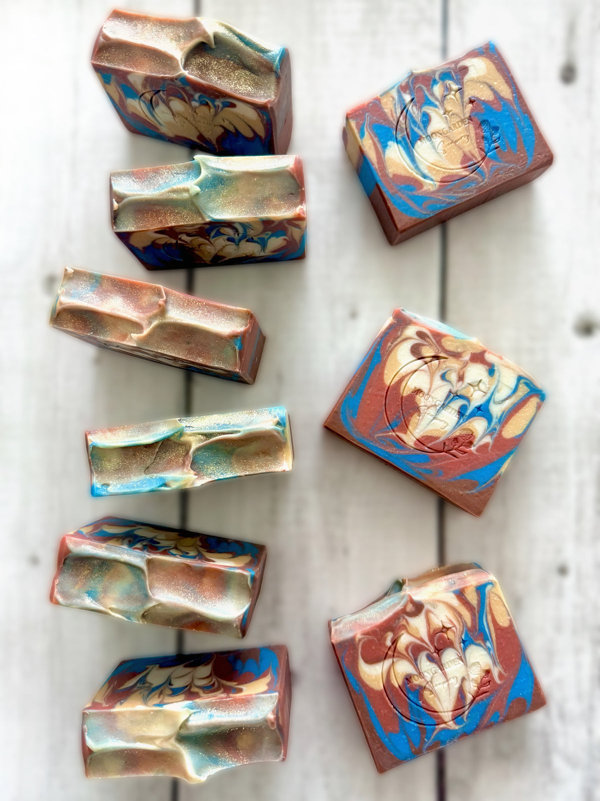 Nine handmade soap bars in blue, cream, brown, white, and sienna, arranged on a white rustic wooden background.