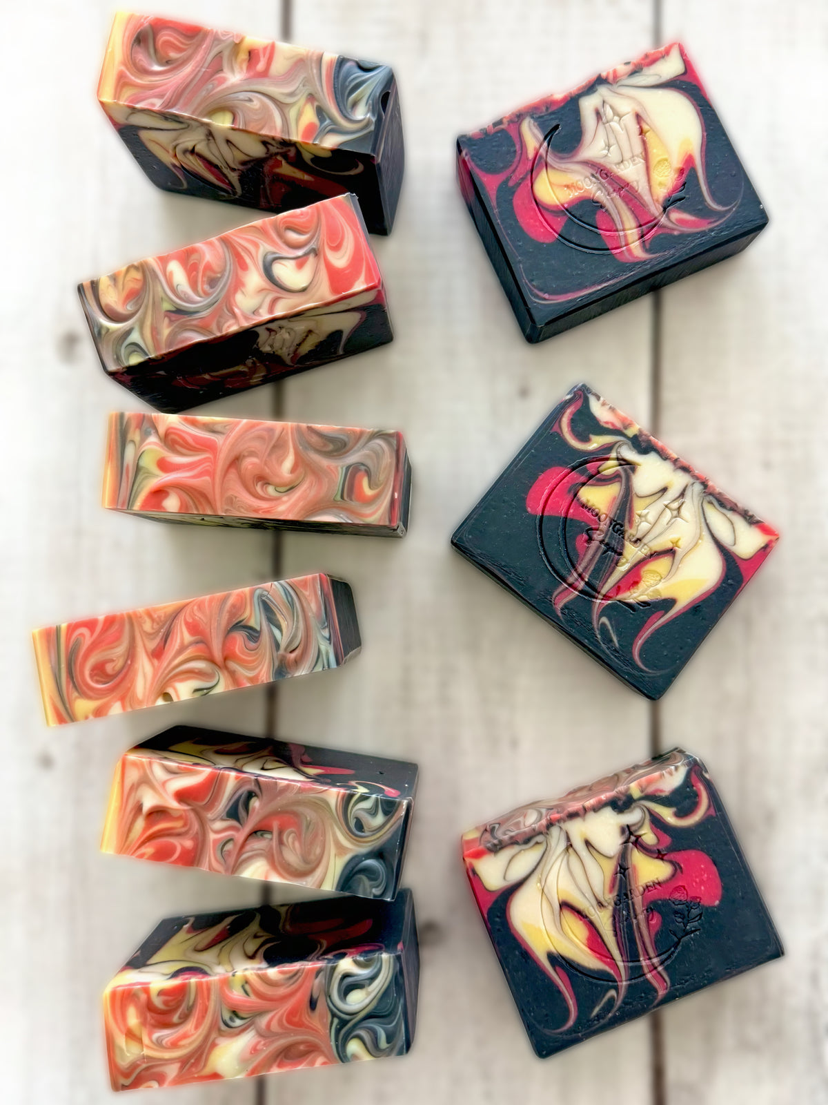Nine handmade soap bars in black, yellow, red, and white, arranged on a white rustic wooden background.