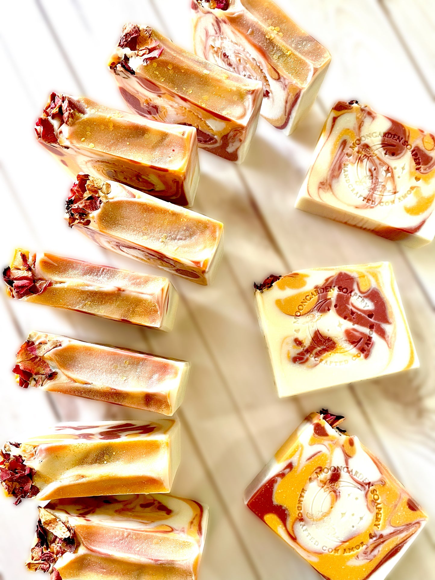 Eleven handmade soap bars in white, red, and gold, topped with glitter, arranged on a white rustic wooden background.