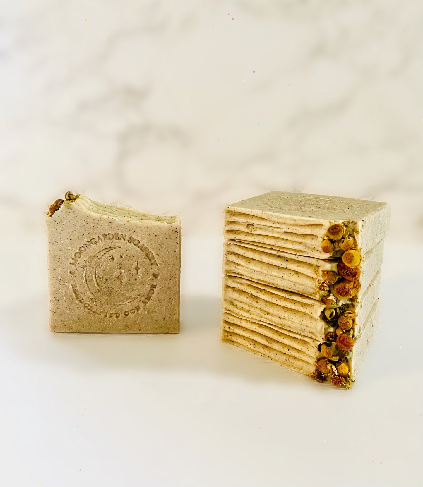 One chamomile and dandelion exfoliating facial bar soap stands on the left. Four chamomile and dandelion exfoliating facial bar soaps are stacked above each other on the right side. Each facial bar soap is topped with dried chamomile flowers on the left. All facial bar soaps sit on top of a marble backdrop.