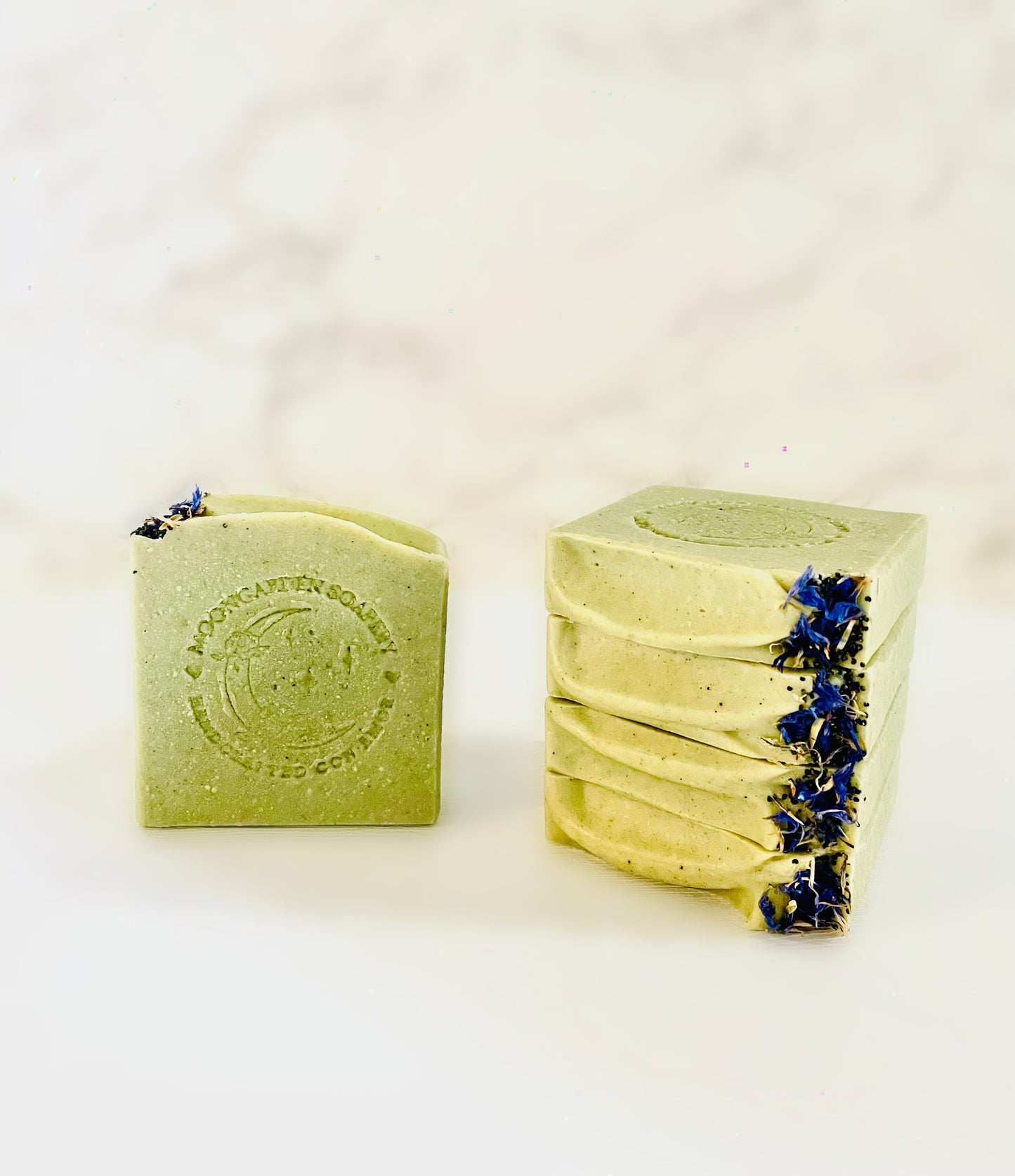 One avocado facial bar soap stands on the left. Four avocado facial bar soaps are stacked above each other on the right side. Each facial bar soap is topped with dried blue cornflowers and poppy seeds on the right. All facial bar soaps sit on top of a marble backdrop.