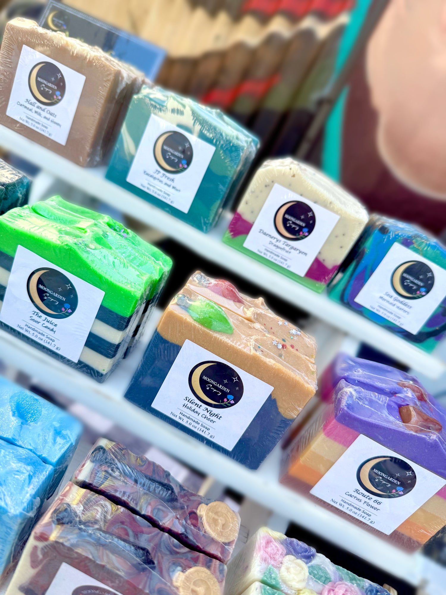 Various bars of handmade soap sitting on a cupcake stand in front of a colorful mural. 