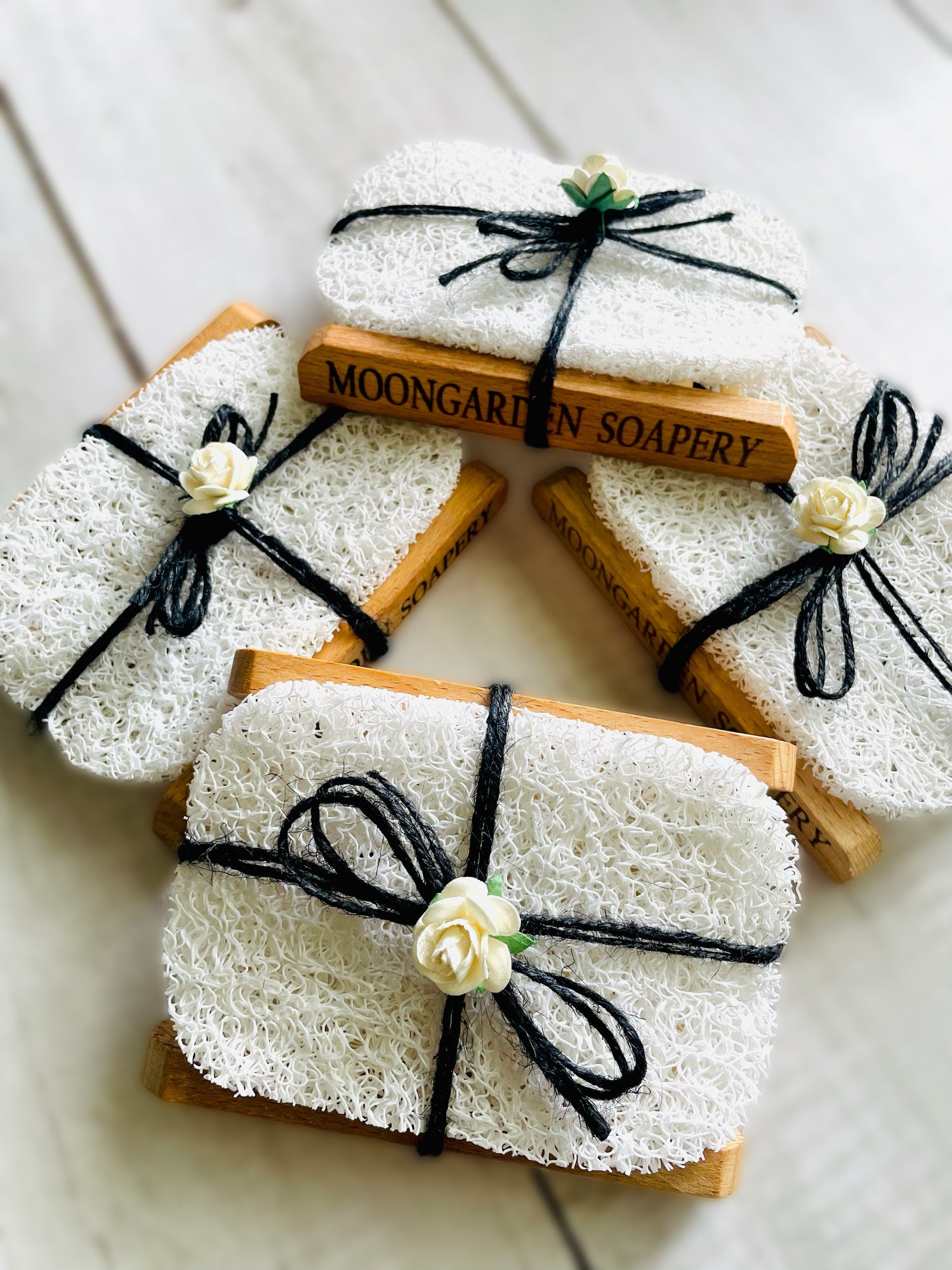 Four soap dishes, each with a soap saver pad tied with black twine string and adorned with a paper rose on top.