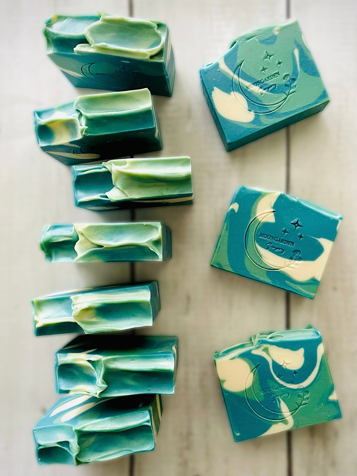 Ten handmade soap bars in light and dark greens with white, arranged on a white rustic wooden background. 