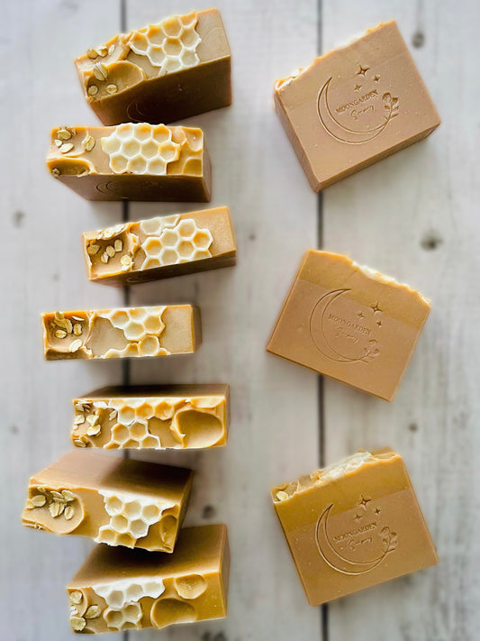 Ten handmade soap bars in tan and gold, topped with oats and honeycomb soap embeds, arranged on a white rustic wooden background. 