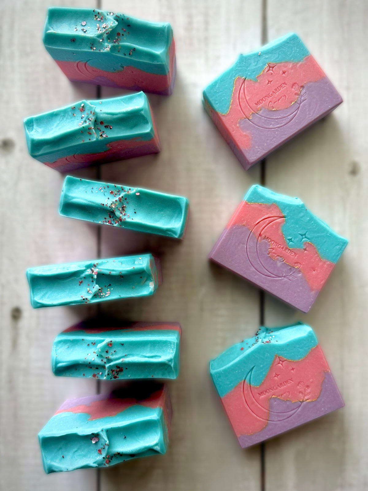 Nine bars of handmade soap bars in light blue, pink, and purple, arranged on a white rustic wooden background. 