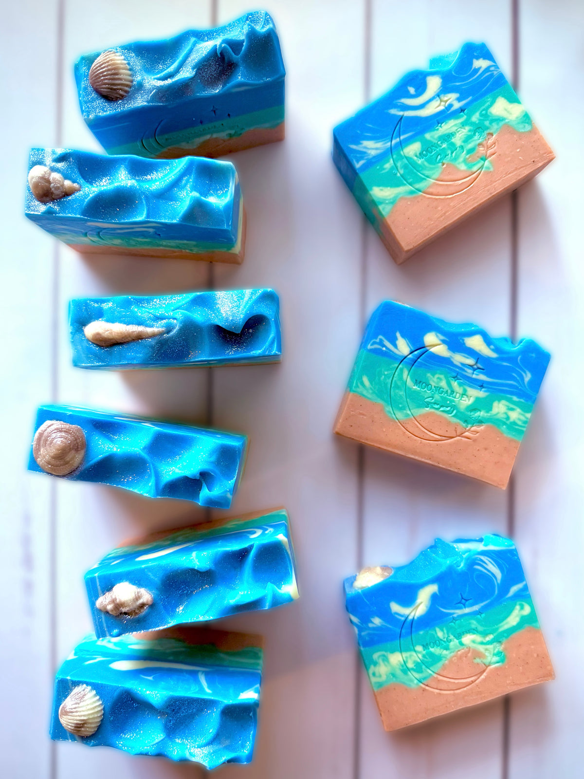 Nine handmade soap bars in blue, light green, white, and tan, topped with seashell soap embeds, arranged on a white rustic wooden background. 