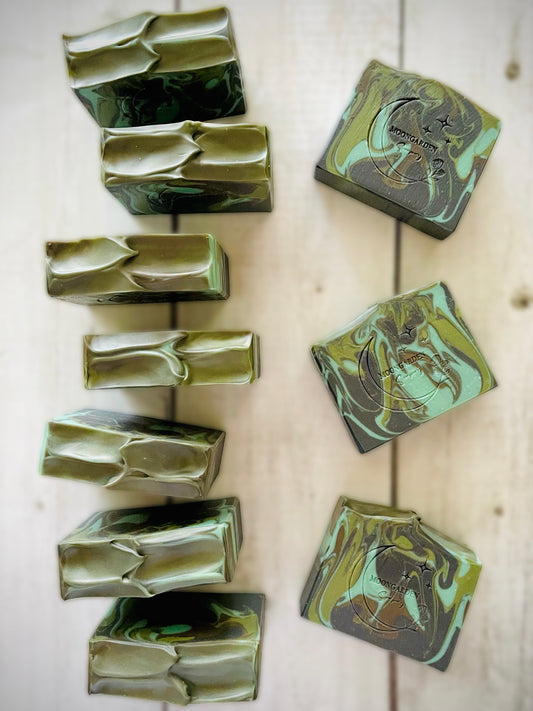 Ten handmade soap bars in light and dark greens and light and dark browns, arranged on a white rustic wooden background. 