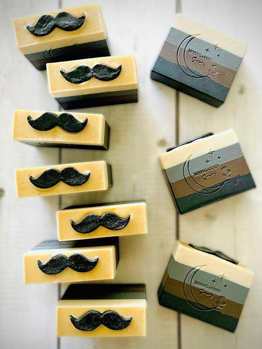 Ten handmade soap bars in black, dark brown, gray, and white, topped with black mustache soap embeds, arranged on a white rustic wooden background. 