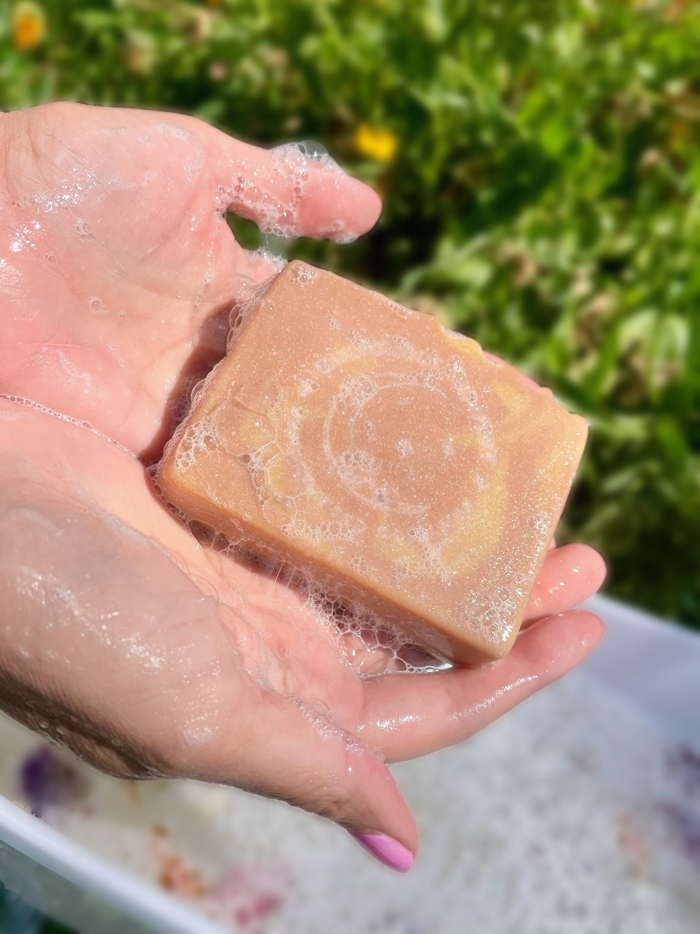 A pair of hands holding bar of soap outside on a sunny day. There is a miniature bath tub below filled with soapy water. There are tiny flowers and grass in the background.