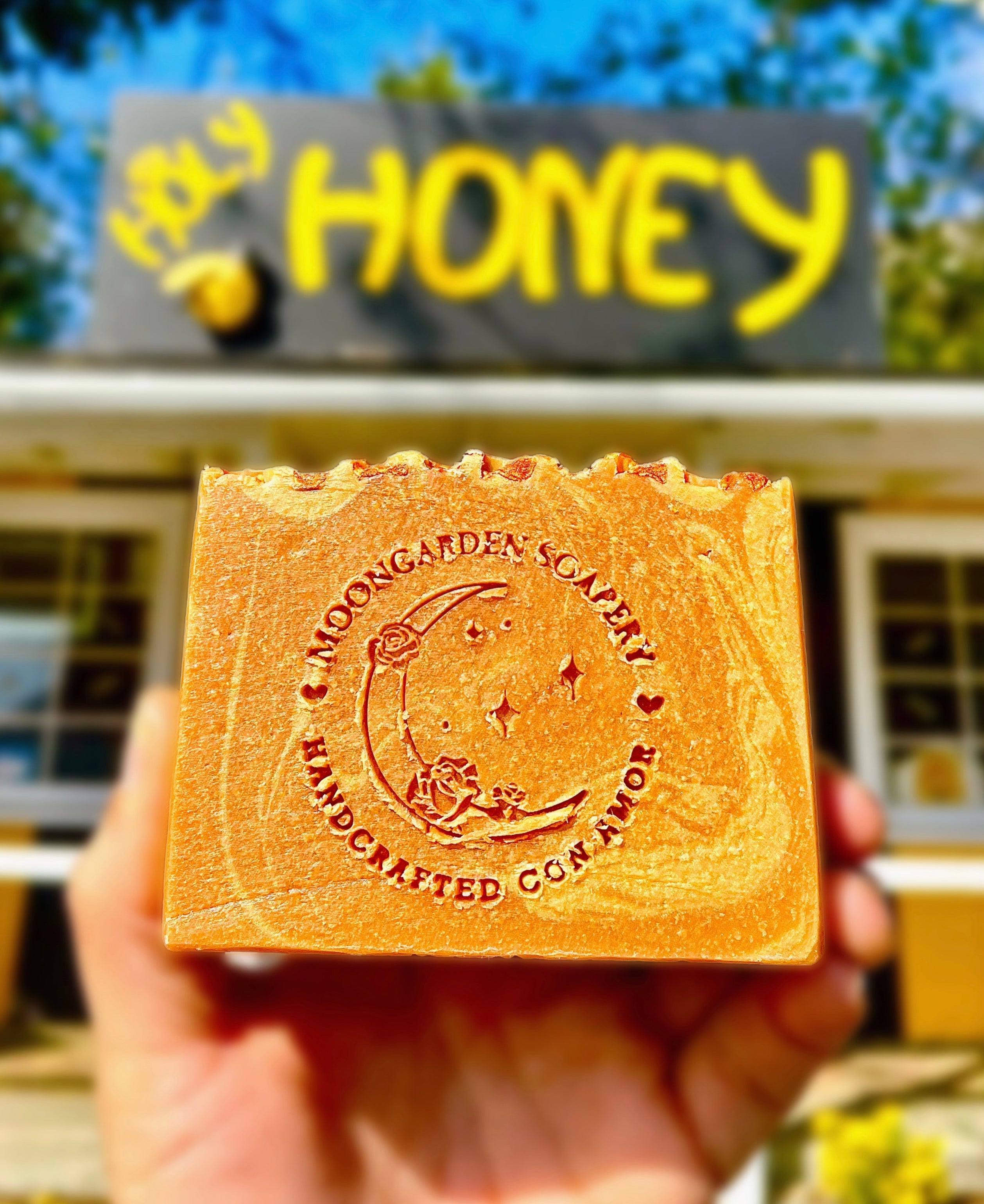 A hand holding a bar of honey soap in front of a honey store. 