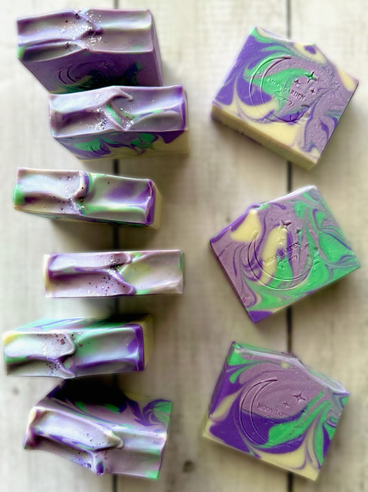 Ten handmade soap bars in light and dark purples, green, and white, topped with glitter, arranged on a white rustic wooden background. 