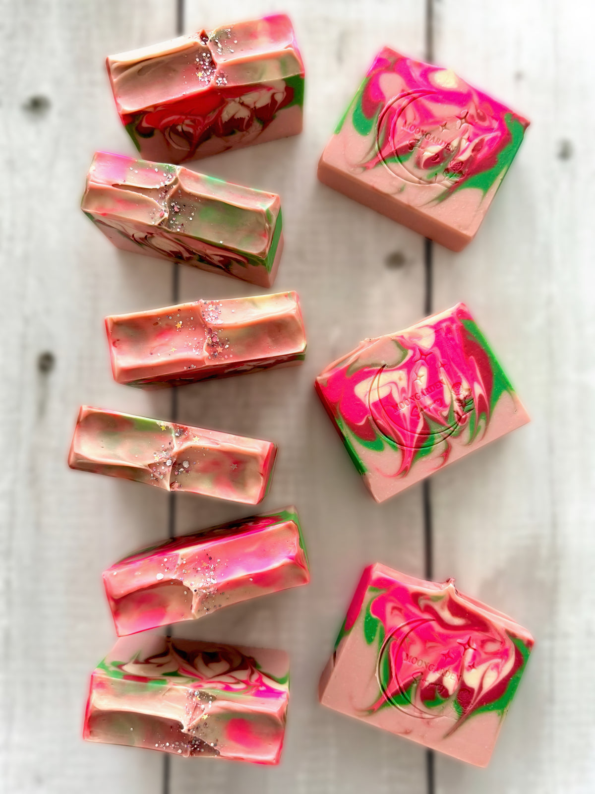 Nine handmade soap bars in neon pink, light pink, green, and white, topped with glitter, arranged on a white rustic wooden background. 