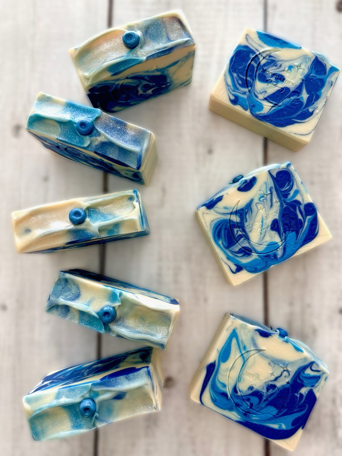 Eight handmade soap bars in white and light and dark blues, topped with blueberry soap embeds, arranged on a white rustic wooden background. 