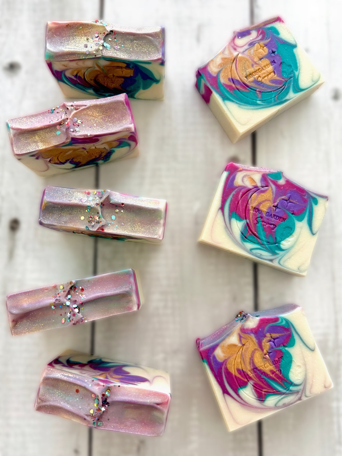 Eight handmade soap bars in purple, pink, sea green, gold, and white, topped with glitter, arranged on a white rustic wooden background. 