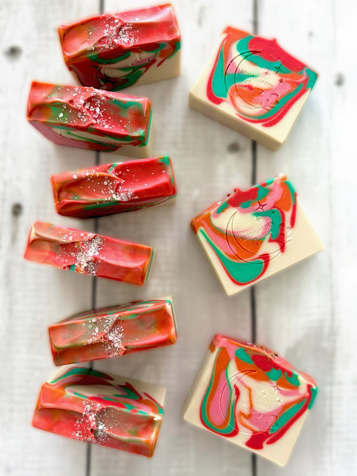Nine handmade soap bars in white, orange, pink, red, and green, topped with glitter, arranged on a white rustic wooden background. 
