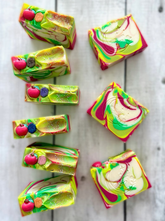 Nine handmade soap bars in dark red, green, white, and bright yellow, topped with glitter, an apple and book soap embed, arranged on a white rustic wooden background. 