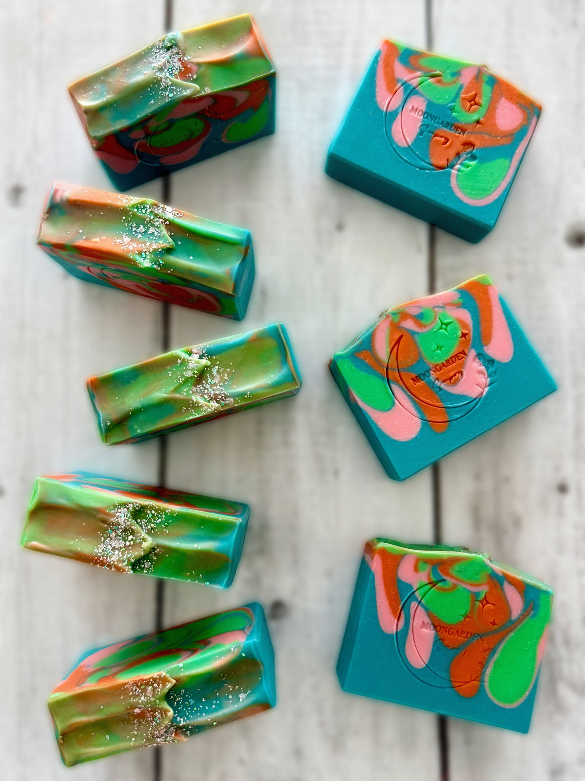 Eight handmade soap bars in sea green, pink, orange, and neon green, topped with glitter, arranged on a white rustic wooden background. 