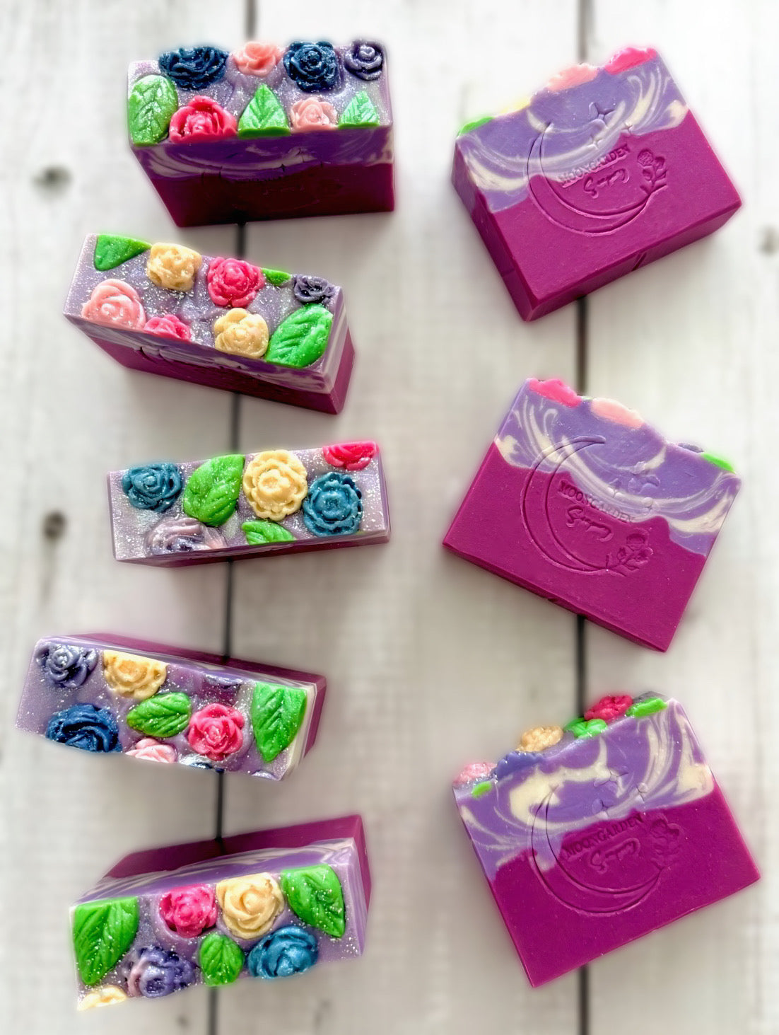 Eight handmade soap bars in dark pink, purple, and white, topped with colorful flower soap embeds, arranged on a white rustic wooden background. 