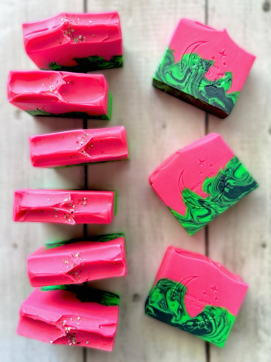 Nine handmade soap bars in neon green, black, and neon pink, topped with glitter, arranged on a white rustic wooden background. 