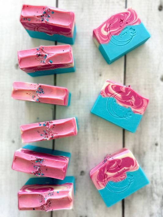 Nine handmade soap bars in  light and dark pinks, light blue, and white, topped with glitter, arranged on a white rustic wooden background. 