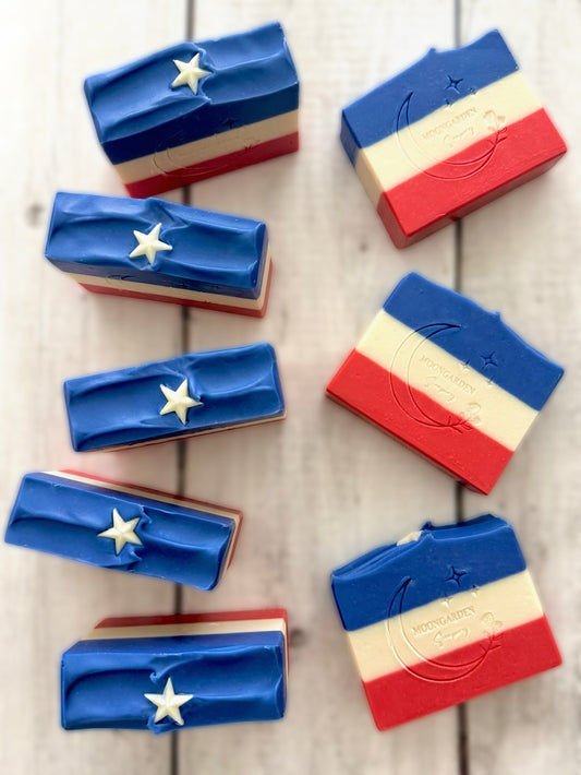 Eight handmade soap bars in red, white, and blue stripes, topped with white star soap embeds, arranged on a white rustic wooden background. 