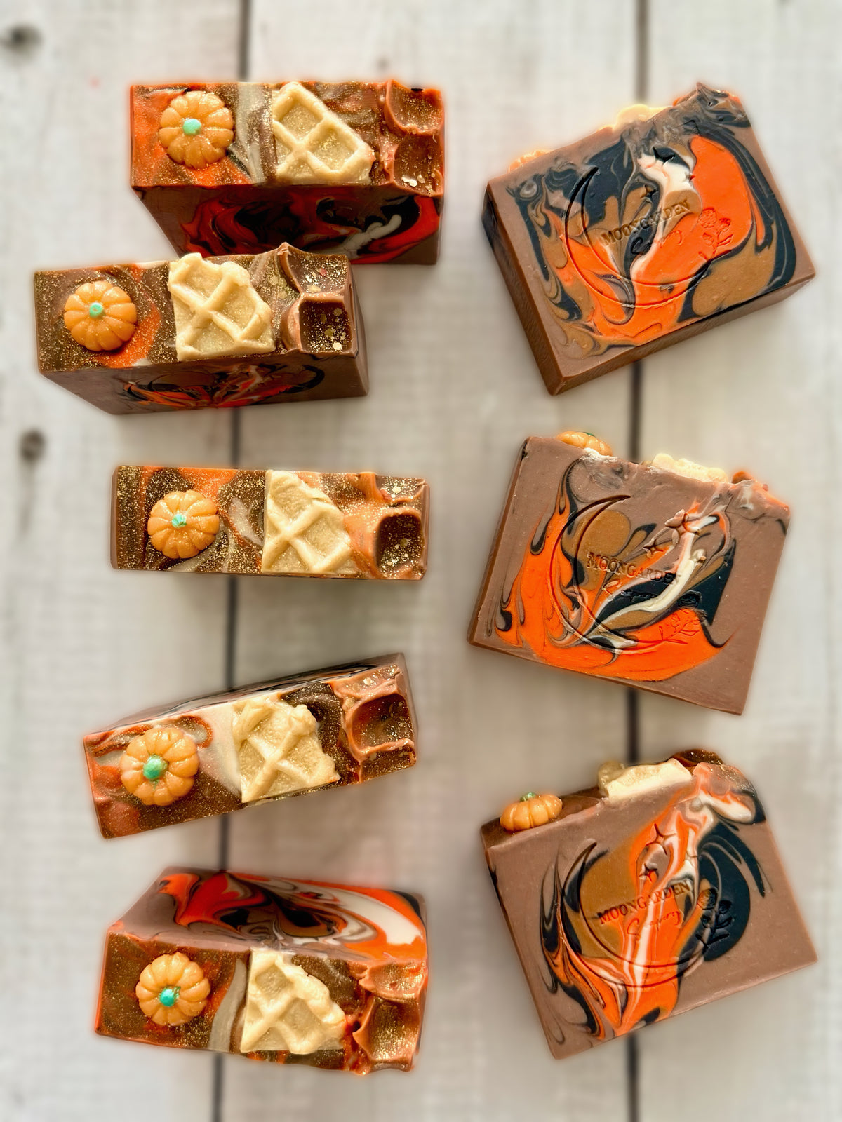 Eight handmade soap bars in tan, brown, orange and white, topped with pumpkin and waffle soap embeds, arranged on a white rustic wooden background. 