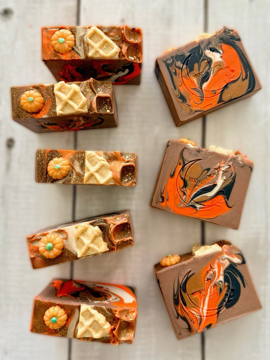 Eight handmade soap bars in tan, brown, orange and white, topped with pumpkin and waffle soap embeds, arranged on a white rustic wooden background. 
