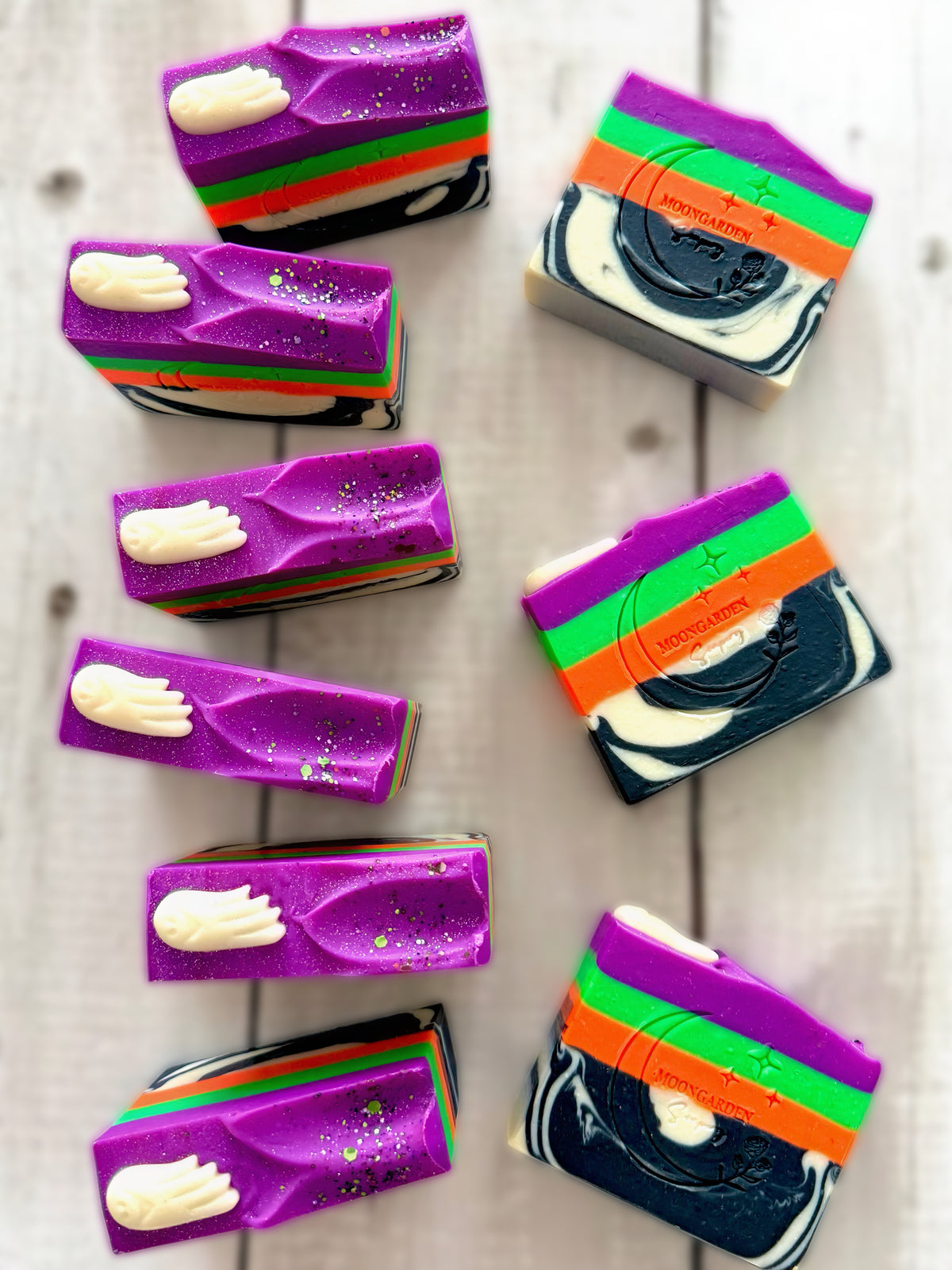 Nine handmade soap bars in black and white with orange, green and purple neons, topped with ghost soap embeds and glitter, arranged on a white rustic wooden background. 