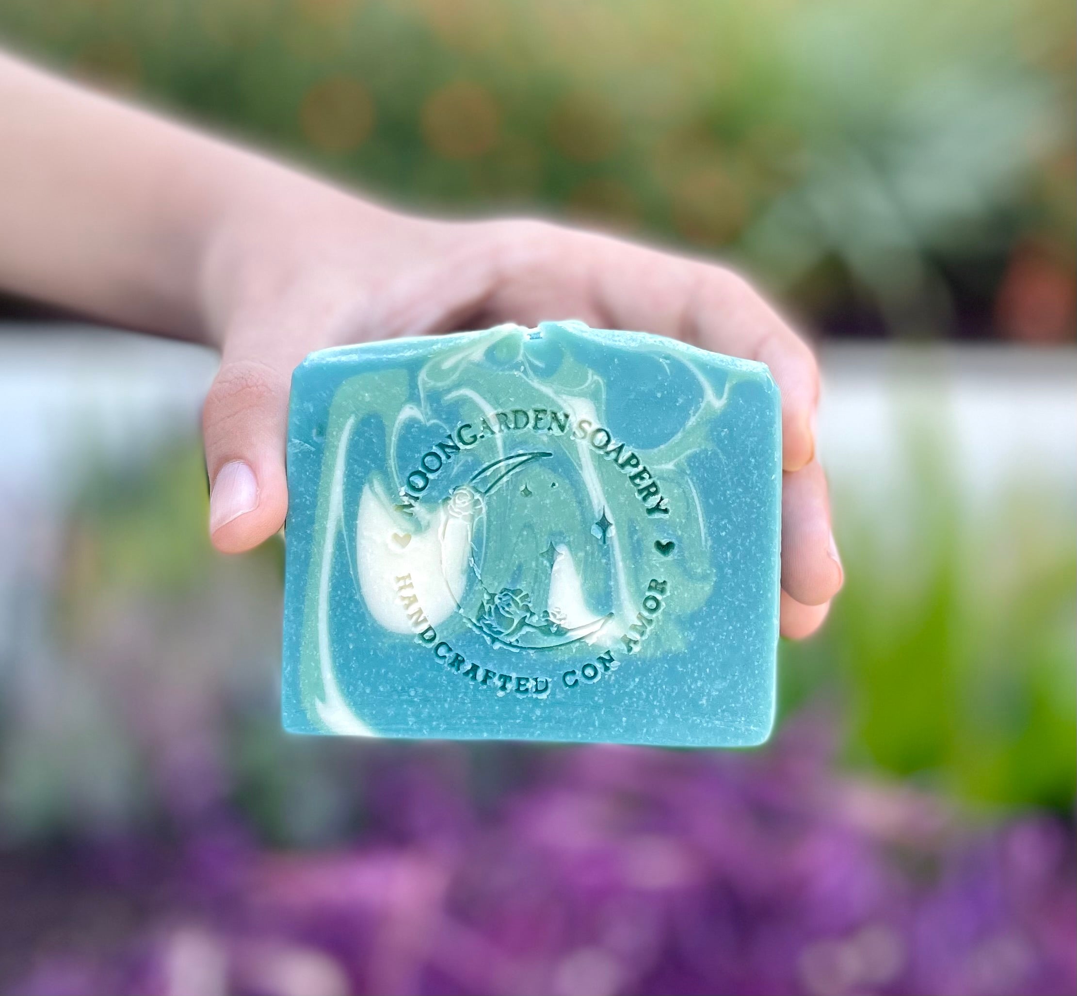 A human hand holding a bar of soap. The background is blurred with flowers and plants. 
