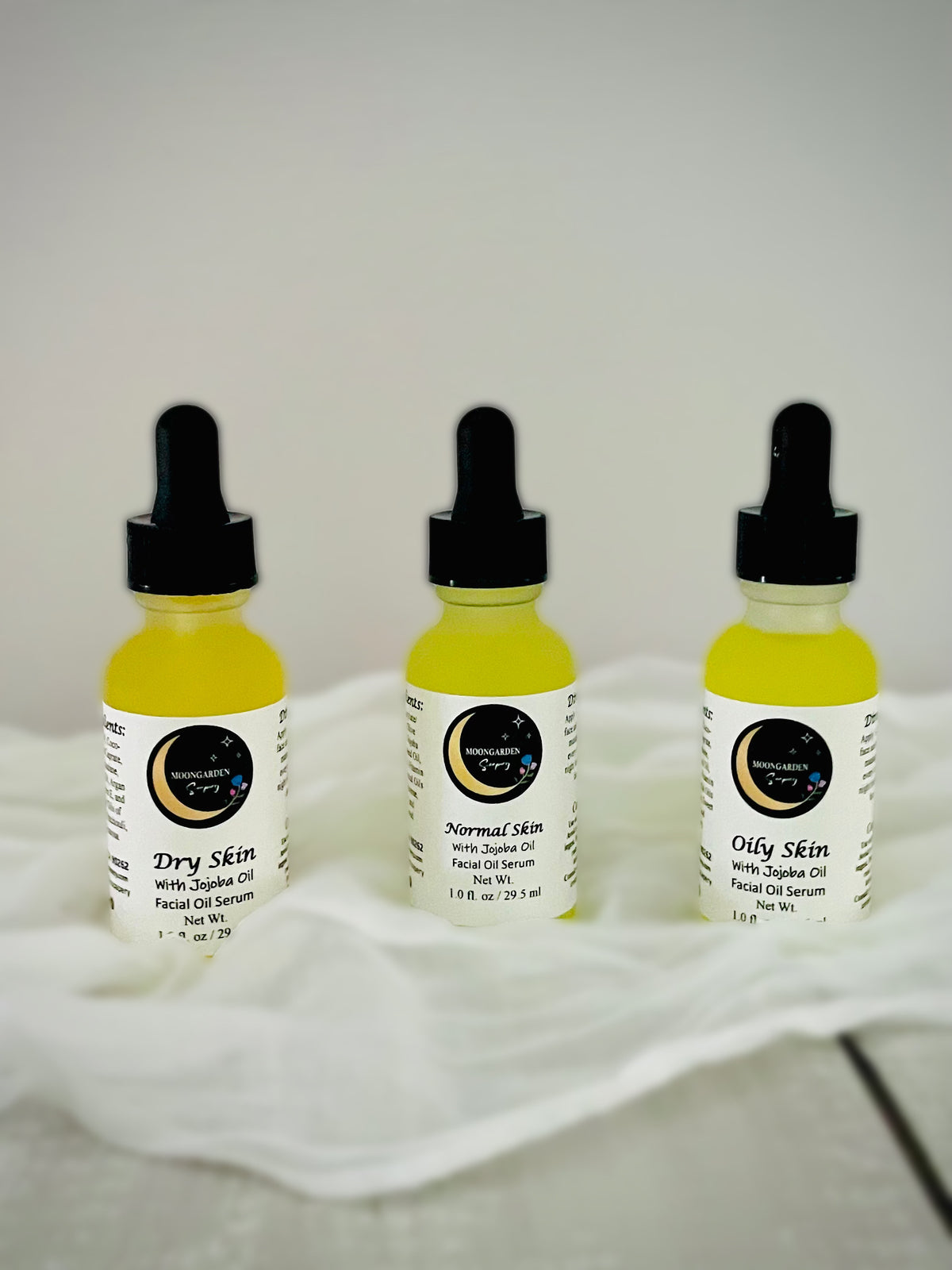 Three small dropper bottles of facial oil arranged on a white fabric surface against a light gray background. 