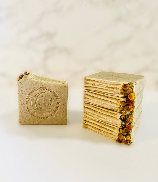 One chamomile and dandelion exfoliating facial bar soap stands on the left. Four chamomile and dandelion exfoliating facial bar soaps are stacked above each other on the right side. Each facial bar soap is topped with dried chamomile flowers on the left. All facial bar soaps sit on top of a marble backdrop. 