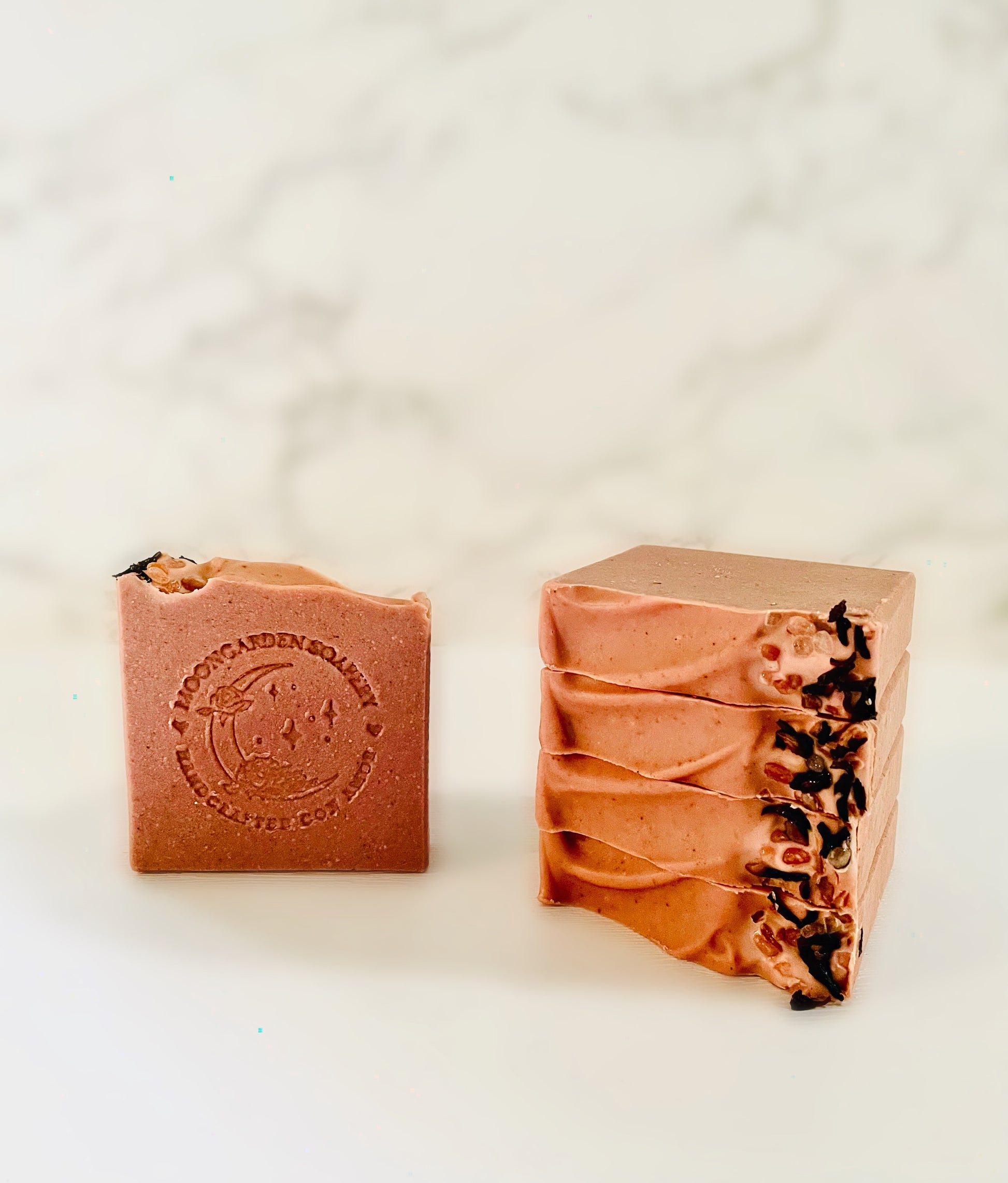 One rose clay facial bar soap stands on the left. Four rose clay facial bar soaps are stacked above each other on the right side. Each facial bar soap is topped with pink Himalayan sea salt and dried hibiscus flowers on the right. All facial bar soaps sit on top of a marble backdrop.