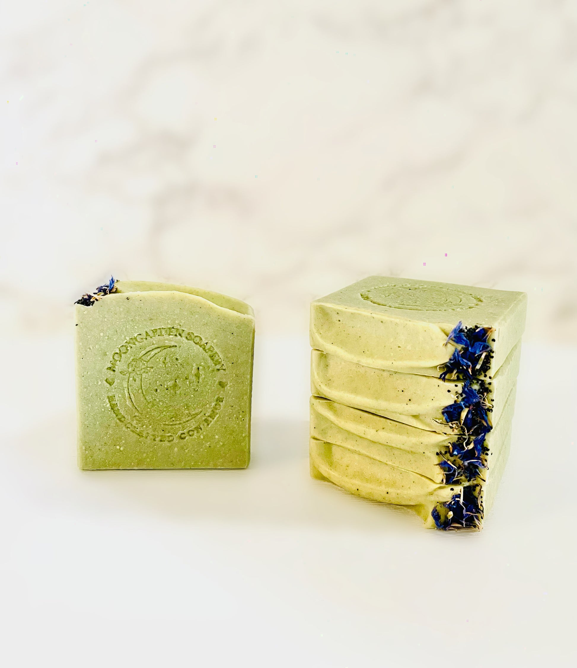 One avocado facial bar soap stands on the left. Four avocado facial bar soaps are stacked above each other on the right side. Each facial bar soap is topped with dried blue cornflowers and poppy seeds on the right. All facial bar soaps sit on top of a marble backdrop. 
