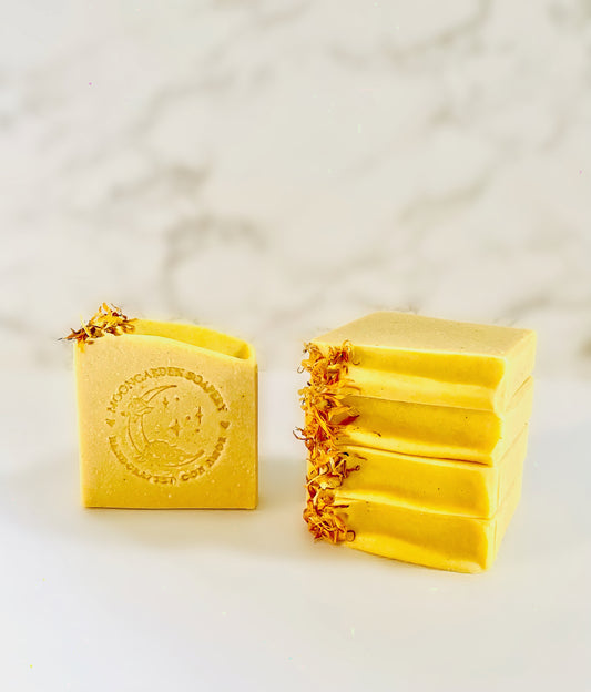 One pumpkin facial bar soap stands on the left. Four pumpkin facial bar soaps are stacked above each other on the right side. Each facial bar soap is topped with dried calendula flowers on the left. All facial bar soaps sit on top of a marble backdrop. 