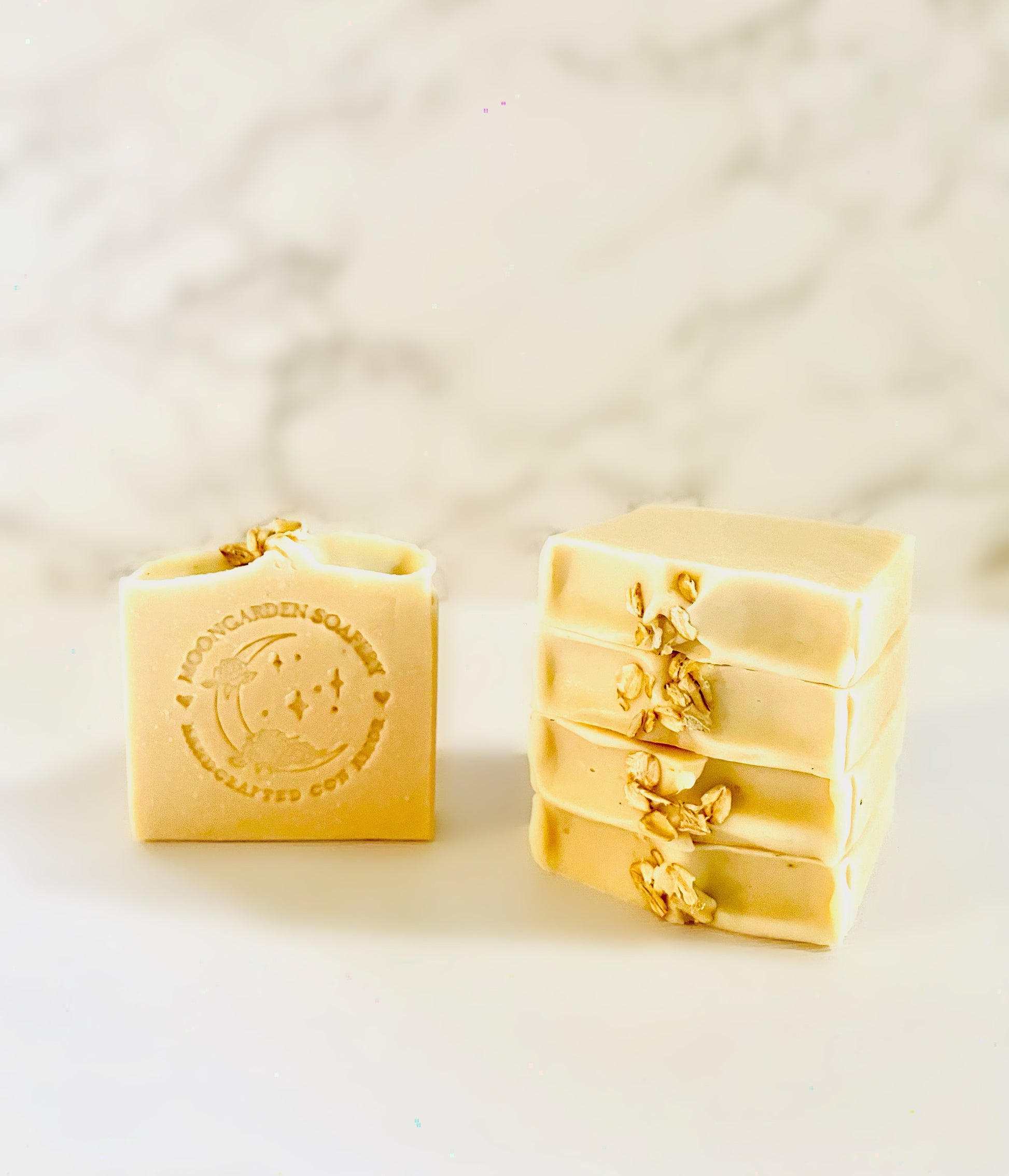 One oats and honey facial bar soap stands on the left. Four oats and honey facial bar soaps are stacked above each other on the right side. Each facial bar soap is topped with whole oats. All facial bar soaps sit on top of a marble backdrop. 