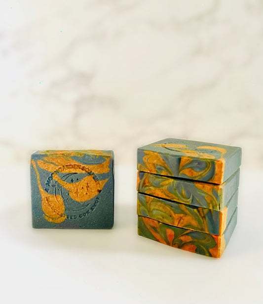 One activated charcoal and turmeric facial bar soap stands on the left. Four activated charcoal and facial bar soaps are stacked above each other on the right side. All facial bar soaps sit on top of a marble backdrop. 